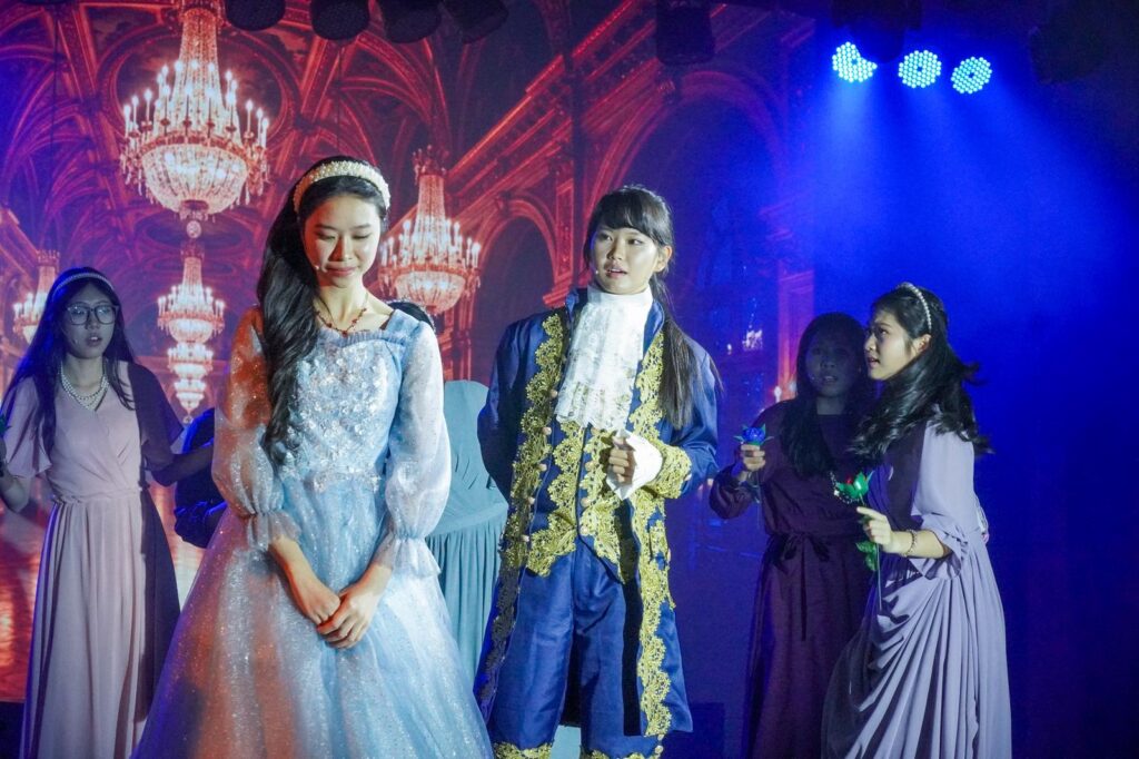 不是每一種學習都能在課室裡完成。有些能力，包括：自信、合作、承擔、面對壓力時的穩定，都需要一個更真實的場景，一群人為同一件事努力，彼此補位，然後在燈光亮起的一刻，把成果交給觀眾。鐘聲慈善社胡陳金枝中學多年來把音樂劇當作校園文化的一部分，並非為了「做一場表演」，而是刻意為學生創造一種獨特的成功感：讓他們在台前台後都能找到位置，感受到自己被需要、能做到、做得到。音樂劇也因此成為一種活動式學習——把語言、情感、價值觀與團隊精神，放進同一個具體而熱烈的練習場。