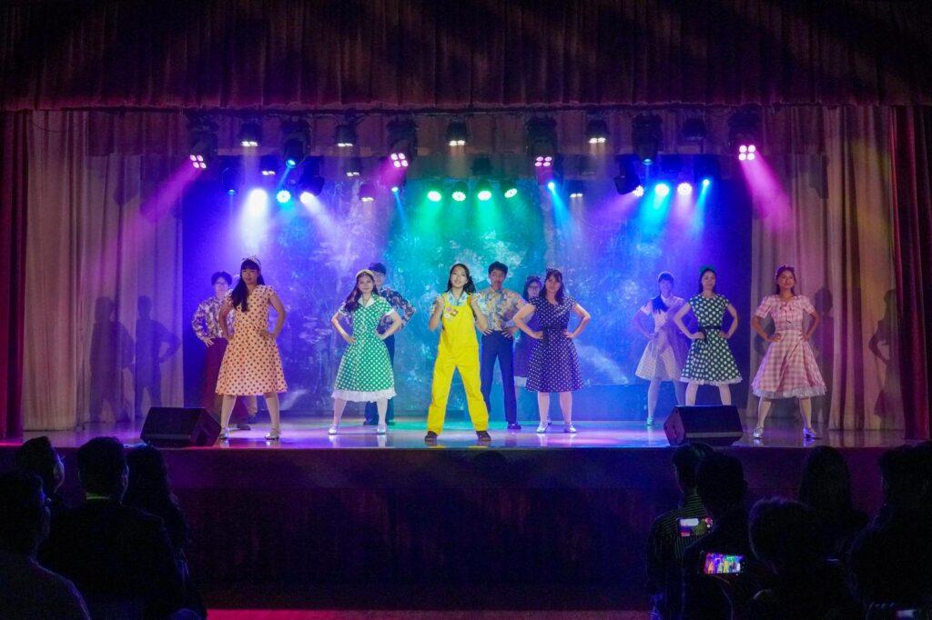 不是每一種學習都能在課室裡完成。有些能力，包括：自信、合作、承擔、面對壓力時的穩定，都需要一個更真實的場景，一群人為同一件事努力，彼此補位，然後在燈光亮起的一刻，把成果交給觀眾。鐘聲慈善社胡陳金枝中學多年來把音樂劇當作校園文化的一部分，並非為了「做一場表演」，而是刻意為學生創造一種獨特的成功感：讓他們在台前台後都能找到位置，感受到自己被需要、能做到、做得到。音樂劇也因此成為一種活動式學習——把語言、情感、價值觀與團隊精神，放進同一個具體而熱烈的練習場。